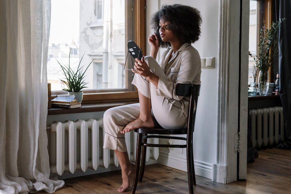Woman in pajamas applying skincare with mirror, enjoying a cozy morning by the window